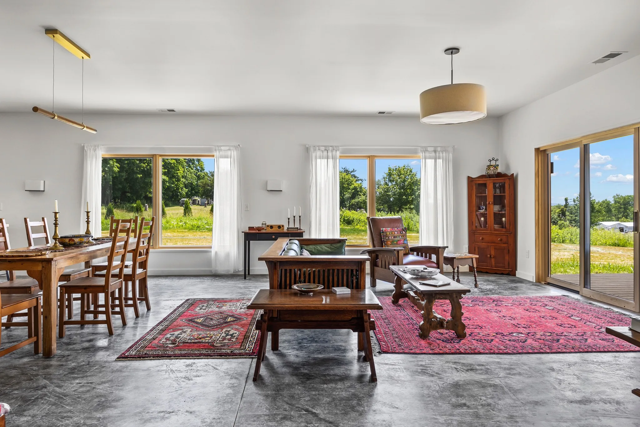 Bright open-concept living room with timber furniture, concrete floors, and large windows overlooking a rural landscape